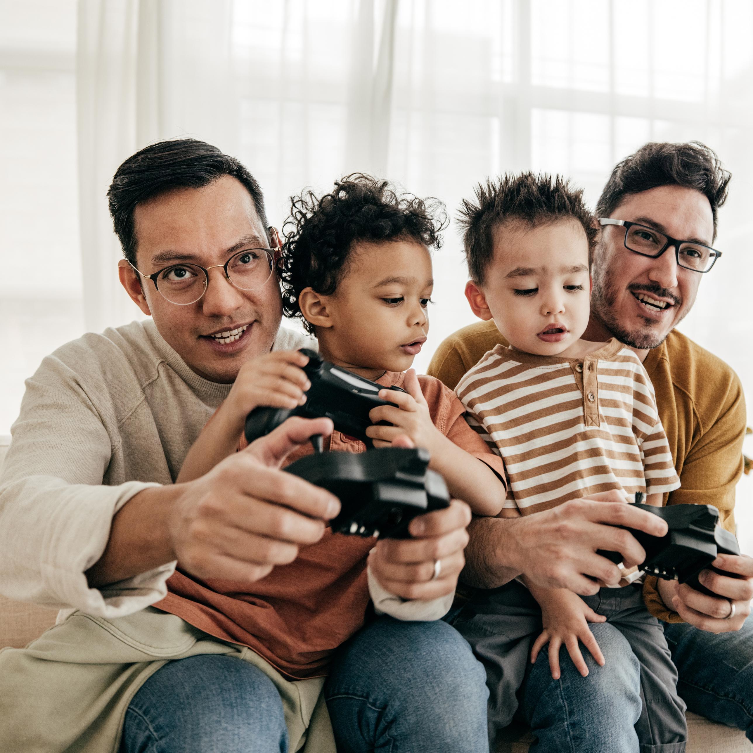 Two queer parents hold their children on their laps teaching them how to play video games.